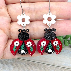 May include: A pair of dangle earrings featuring a white flower stud connected to a ladybug charm. The ladybug has a black glitter head and red wings decorated with white flowers and green leaves. The earrings hang from silver-toned metal hooks.