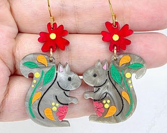 May include: A pair of dangle earrings featuring gray squirrel-shaped charms with marbled textures. Each squirrel is decorated with painted green leaves, orange teardrop shapes, and yellow dots. A bright red flower with a yellow center sits above each squirrel, attached to a gold-toned metal hook.