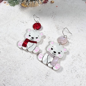May include: A pair of whimsical bear earrings. Each earring features a white bear with a red or pink scarf, pink paws, and a red or pink accent above. The earrings have silver-colored hooks.