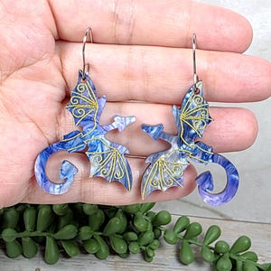 May include: A pair of dragon-shaped earrings. The earrings feature a blue and white marbled design with gold-colored filigree accents on the wings. The dragon's body has a curled tail. The earrings are suspended from silver-colored hooks.