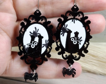 May include: A pair of drop earrings featuring an oval black frame with intricate scrollwork detailing. The centre displays a white background with a black cat silhouette and spider graphics. A small black spider charm hangs from each earring.