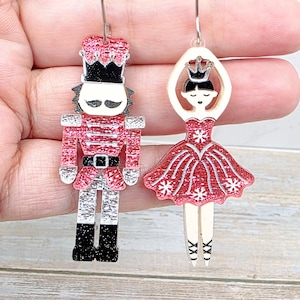 May include: A pair of mismatched dangle earrings featuring a nutcracker and a ballerina. The nutcracker has a red glitter coat, black boots, and a hat. The ballerina wears a red glitter tutu with white snowflake details and a crown.