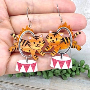 May include: A pair of dangle earrings featuring orange tigers with black stripes jumping through silver hoops atop white circus pedestals with red triangular accents. The hoops are topped with orange glittery flames, and the earrings hang from silver hooks.