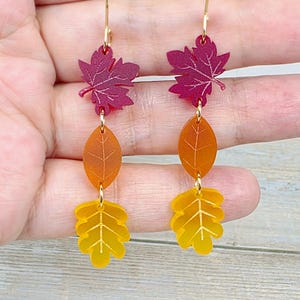 May include: A pair of dangle earrings featuring three stacked leaf-shaped charms in descending order. The top charm is a deep magenta maple leaf, the middle is an orange oval leaf, and the bottom is a yellow oak leaf, all on gold-tone hooks.