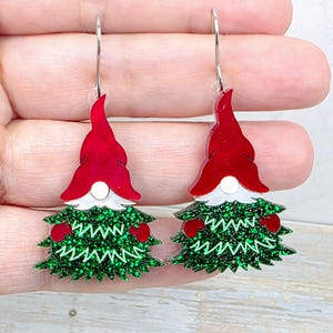 May include: A pair of dangle earrings featuring a gnome design with a shiny red hat, a white beard, and a green glittery body. The earrings are suspended from silver-toned metal hooks and showcase a festive, layered appearance with small red accents on the sides.