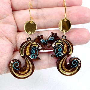May include: A pair of dangle earrings featuring brown squirrel-shaped charms with gold glitter accents, teal swirl patterns, and small white dots. Each squirrel hangs from a gold-toned circular connector attached to a gold-toned metal hook wire.