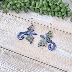 May include: A pair of dragon-shaped earrings. The earrings feature a blue and purple marbled design with gold filigree wings and accents. The earrings hang from silver-colored ear wires. The earrings are displayed on a light wood surface.