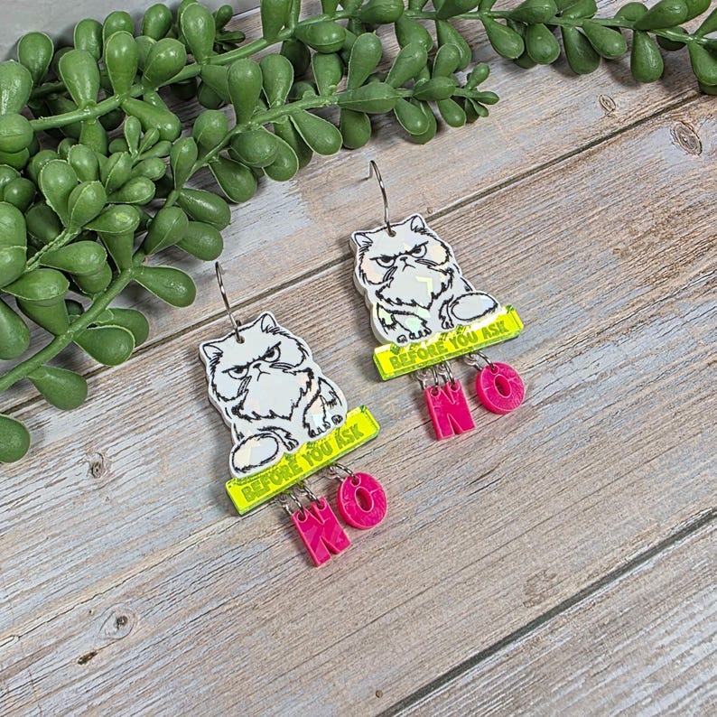 May include: A pair of novelty earrings featuring a grumpy white cat illustration. Each earring has a yellow banner with the text "BEFORE YOU ASK" and the word "NO" in pink. The earrings hang from silver-colored hooks.