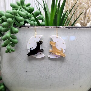 May include: A pair of dangle earrings featuring white crescent moon charms with gold star accents. One earring displays a black dog silhouette, while the other features a gold-toned dog silhouette, both suspended from gold-colored metal hooks.
