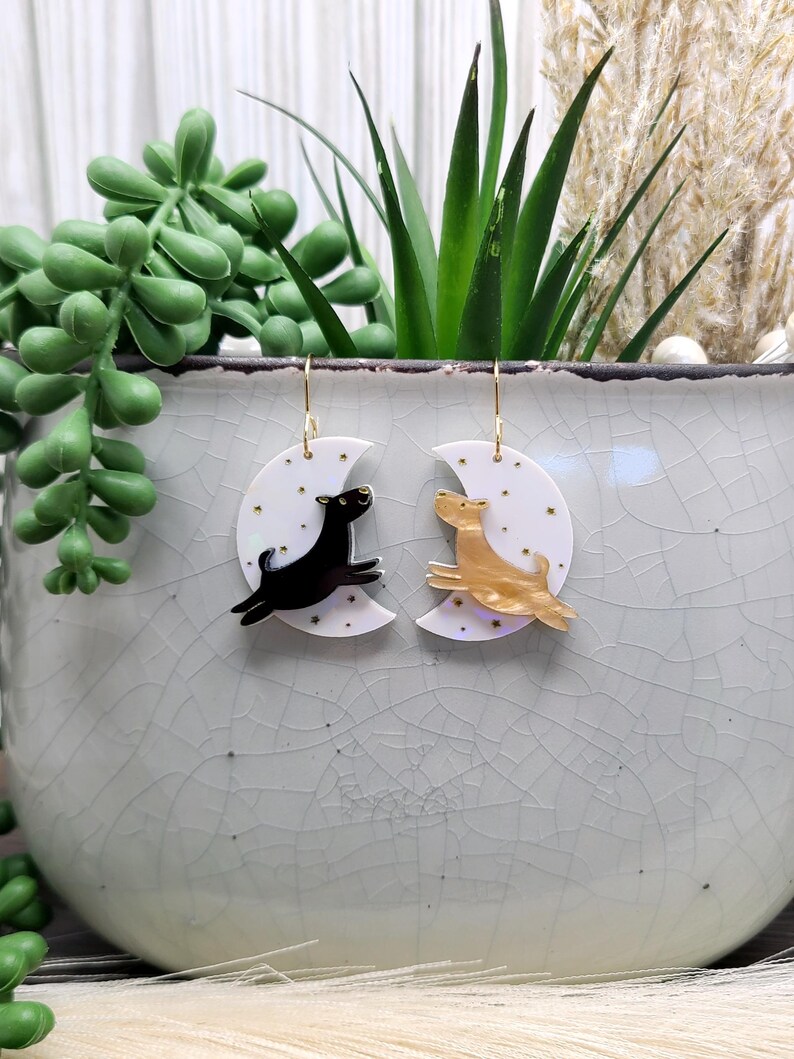 May include: A pair of dangle earrings featuring white crescent moon charms with gold star accents. One earring displays a black dog silhouette, while the other features a gold-toned dog silhouette, both suspended from gold-colored metal hooks.