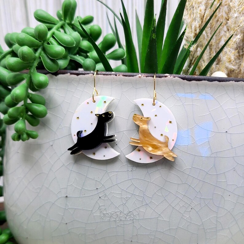 May include: A pair of dangle earrings featuring white crescent moon charms with iridescent finishes and small gold star accents. One earring displays a black dog silhouette, while the other features a tan marbled dog silhouette, both suspended from gold-toned hooks.