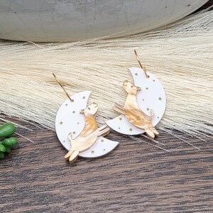 May include: A pair of dangle earrings featuring a white crescent moon shape with small gold star accents. A marbled tan and cream-colored dog figure is layered over each moon. The earrings are attached to gold-toned metal hooks for hanging.