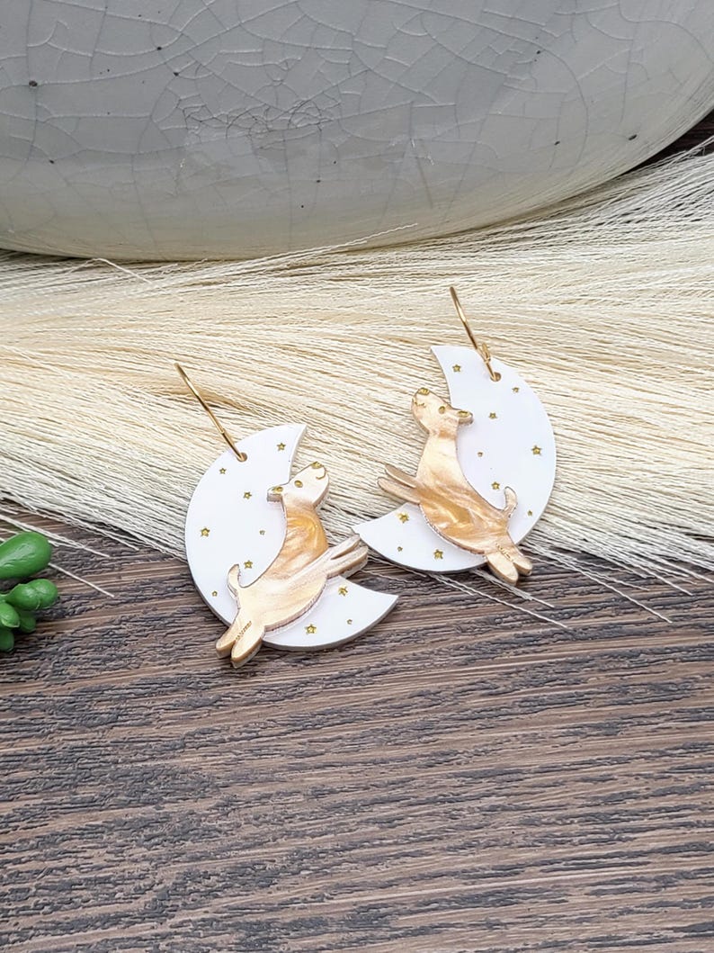 May include: A pair of dangle earrings featuring a white crescent moon shape with small gold star accents. A marbled tan and cream-colored dog figure is layered over each moon. The earrings are attached to gold-toned metal hooks for hanging.