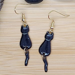 May include: A pair of black cat earrings with gold-colored hooks. The earrings feature a glossy black cat silhouette with a white accent line and a dangling tail. The earrings are displayed on a light brown wooden surface.