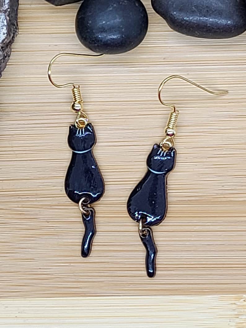 May include: A pair of black cat earrings with gold-colored hooks. The earrings feature a glossy black cat silhouette with a white accent line and a dangling tail. The earrings are displayed on a light brown wooden surface.