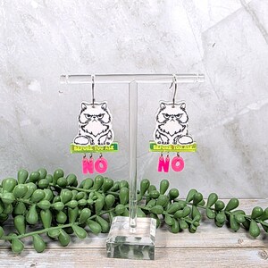 May include: A pair of acrylic earrings featuring a grumpy cat design. Each earring has a white and black cat image above a green rectangle with the text "BEFORE YOU ASK" and the pink word "NO". The earrings hang from silver-colored hooks.
