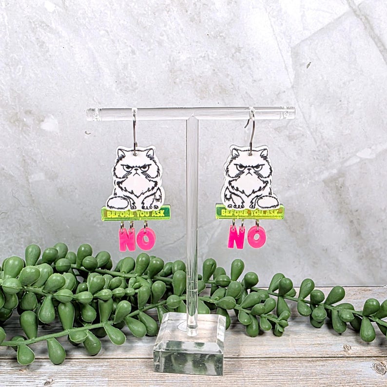 May include: A pair of acrylic earrings featuring a grumpy cat design. Each earring has a white and black cat image above a green rectangle with the text "BEFORE YOU ASK" and the pink word "NO". The earrings hang from silver-colored hooks.