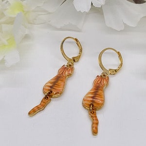 May include: A pair of gold-colored leverback earrings. Each earring features a detailed orange and brown tabby cat design with a dangling tail. The earrings are set against a soft, white background.