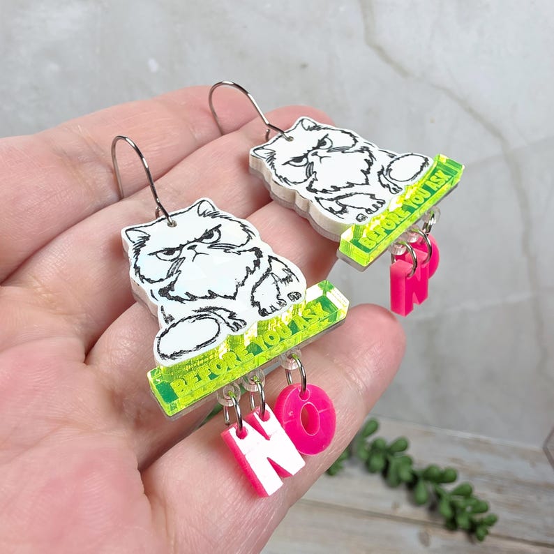 May include: A pair of novelty earrings featuring a grumpy white cat illustration with the text "Before You Ask" on a green bar. Below, pink letters spell "NO". The earrings have silver-colored hooks.