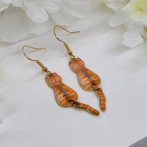 May include: A pair of gold-colored cat earrings. Each earring features a detailed orange and brown tabby cat design with a dangling tail. The earrings have a hook closure.