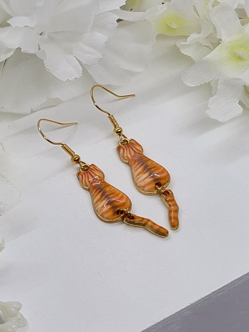 May include: A pair of gold-colored cat earrings. Each earring features a detailed orange and brown tabby cat design with a dangling tail. The earrings have a hook closure.