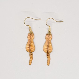May include: A pair of gold-colored earrings featuring a cat's rear end design. The earrings have a tan and orange striped pattern, with gold-colored ear hooks. The earrings are designed to dangle.