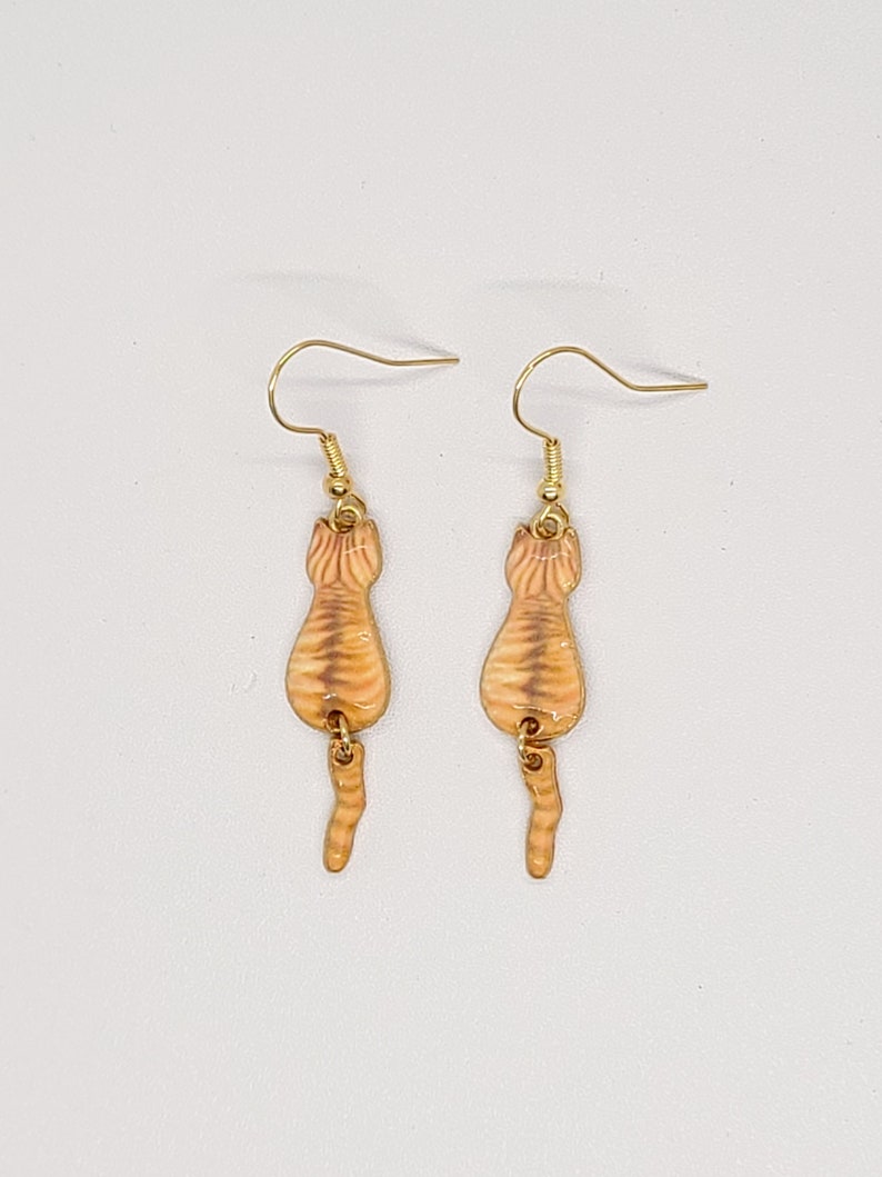 May include: A pair of gold-colored earrings featuring a cat's rear end design. The earrings have a tan and orange striped pattern, with gold-colored ear hooks. The earrings are designed to dangle.