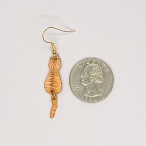 May include: A single gold-colored earring featuring a small, orange tabby cat design. The cat is depicted from the rear, with visible stripes and a curled tail. The earring is shown next to a U.S. quarter for size comparison.