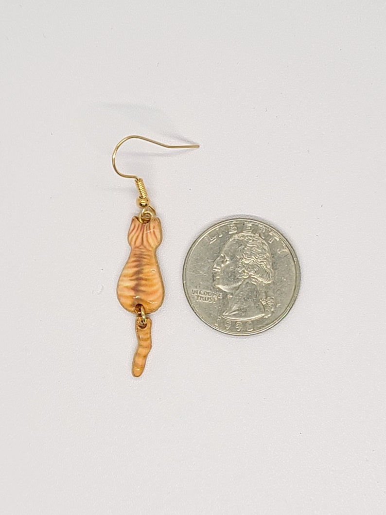 May include: A single gold-colored earring featuring a small, orange tabby cat design. The cat is depicted from the rear, with visible stripes and a curled tail. The earring is shown next to a U.S. quarter for size comparison.