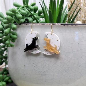 May include: A pair of dangle earrings featuring white crescent moon charms decorated with small gold stars. One earring displays a black dog silhouette leaping across the moon, while the other shows a gold-toned dog silhouette in a similar pose.