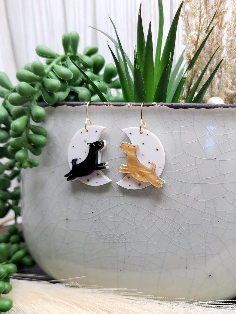 May include: A pair of dangle earrings featuring white crescent moon charms decorated with small gold stars. One earring displays a black dog silhouette leaping across the moon, while the other shows a gold-toned dog silhouette in a similar pose.