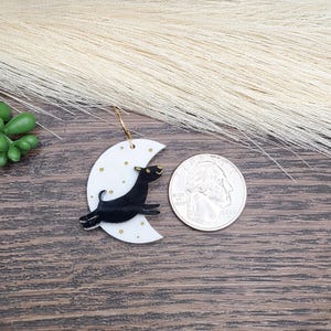 May include: A white crescent moon earring featuring a black dog leaping across the surface, accented with small gold stars. The earring is displayed next to a United States quarter coin for scale, showing the relative size of the jewelry piece.