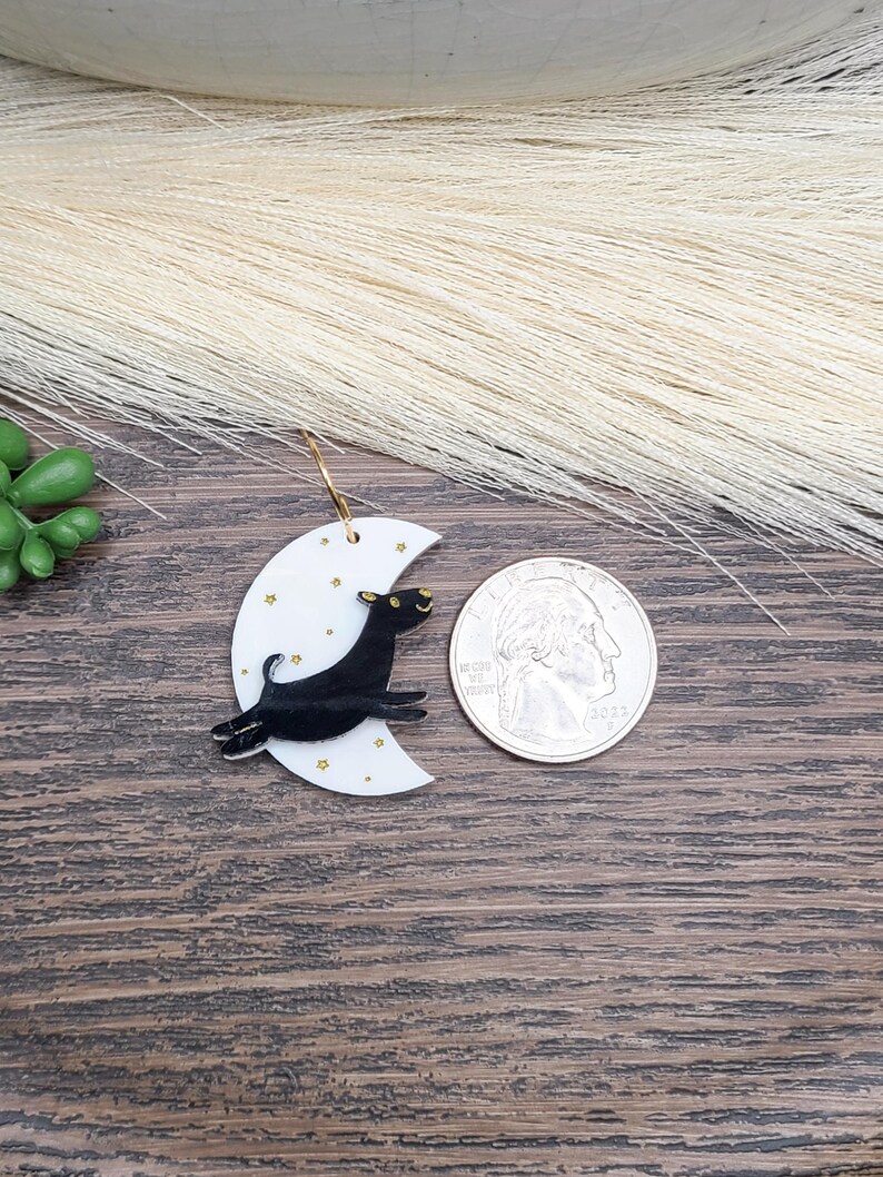 May include: A white crescent moon earring featuring a black dog leaping across the surface, accented with small gold stars. The earring is displayed next to a United States quarter coin for scale, showing the relative size of the jewelry piece.