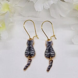 May include: A pair of gold-tone cat earrings. Each earring features a gray and white striped cat body with a dangling tail. The earrings have a hook closure.