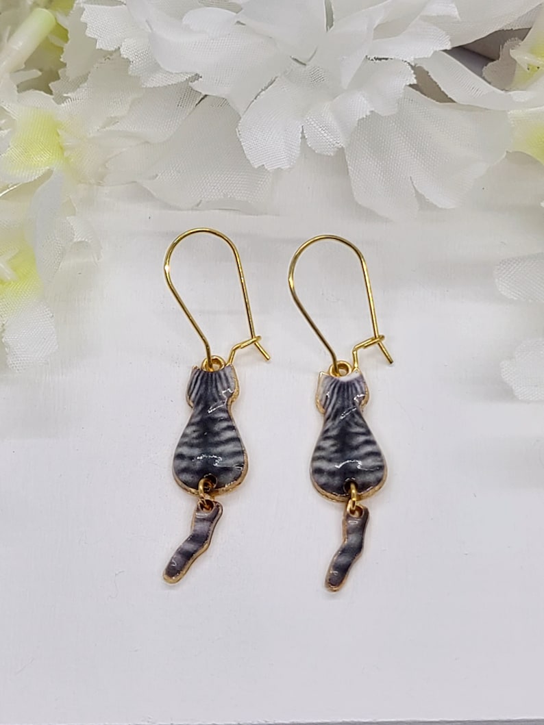 May include: A pair of gold-tone cat earrings. Each earring features a gray and white striped cat body with a dangling tail. The earrings have a hook closure.