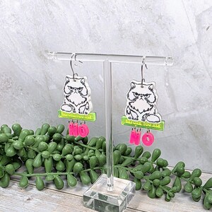 May include: A pair of dangle earrings featuring a grumpy cat design. Each earring has a white cat illustration with the text "BEFORE YOU ASK" above and the word "NO" in pink below. The earrings hang from silver-colored hooks.