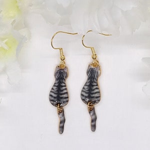 May include: A pair of gold-tone cat earrings. Each earring features a gray and black striped cat design with a dangling tail. The earrings are suspended from gold-colored ear wires.