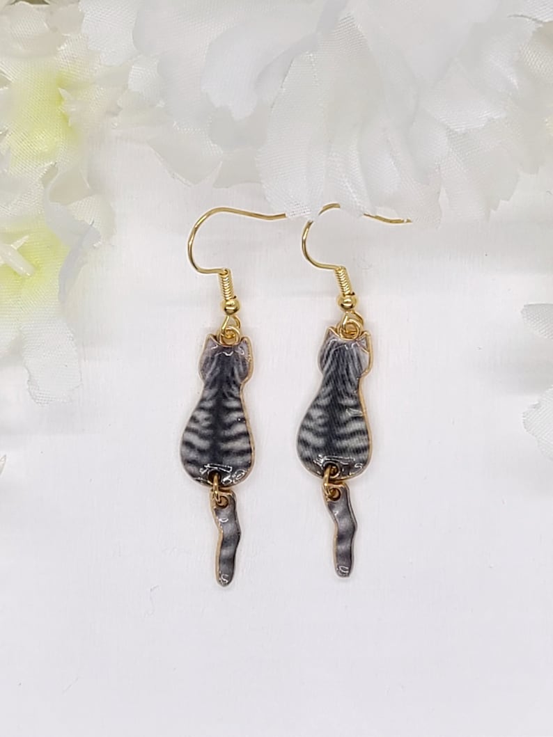 May include: A pair of gold-tone cat earrings. Each earring features a gray and black striped cat design with a dangling tail. The earrings are suspended from gold-colored ear wires.