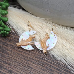 May include: A pair of dangle earrings featuring iridescent white crescent moons with small gold star accents. Each moon is adorned with a gold-toned dog figure in a jumping pose. The earrings are suspended from thin gold-colored metal hooks.