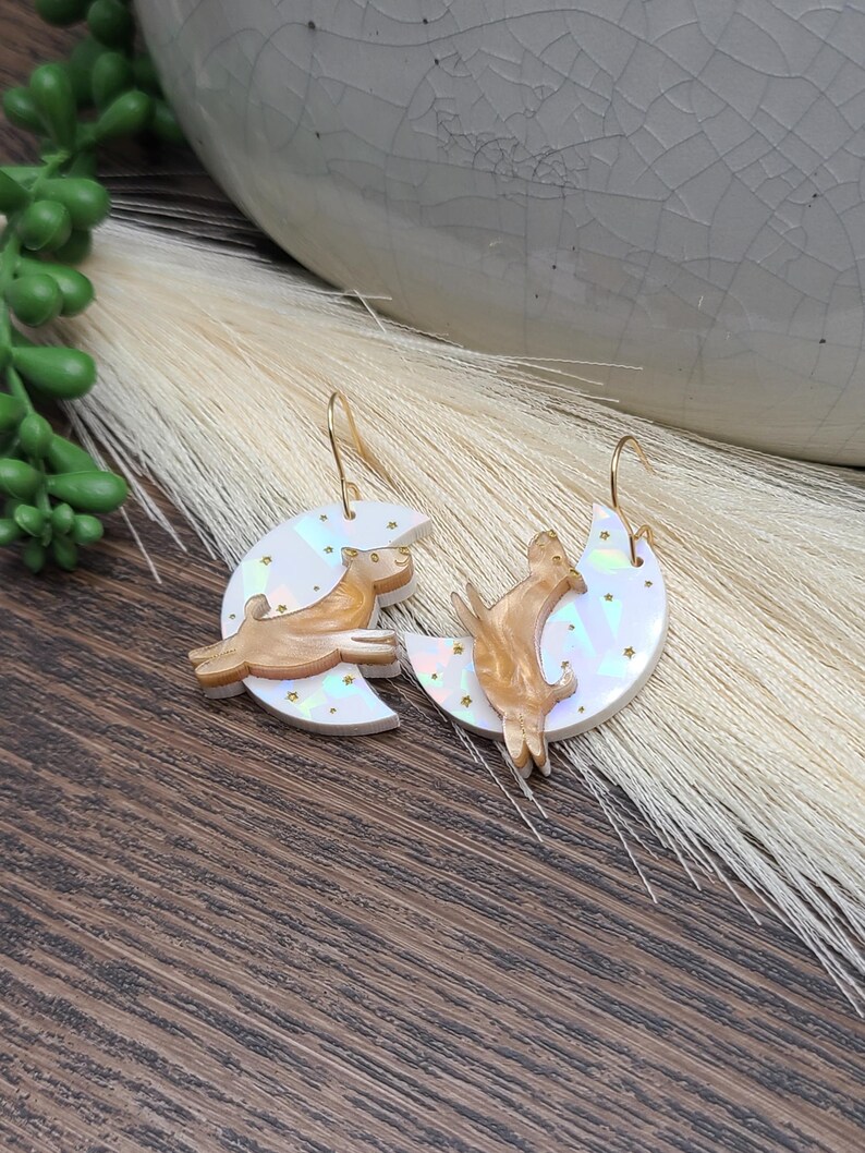 May include: A pair of dangle earrings featuring iridescent white crescent moons with small gold star accents. Each moon is adorned with a gold-toned dog figure in a jumping pose. The earrings are suspended from thin gold-colored metal hooks.