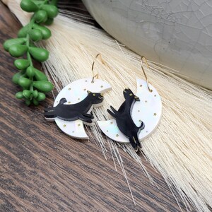 May include: A pair of dangle earrings featuring a white crescent moon with a subtle iridescent finish and small gold star accents. A black dog silhouette is layered on top of each moon, and the earrings hang from gold-toned metal ear wires.