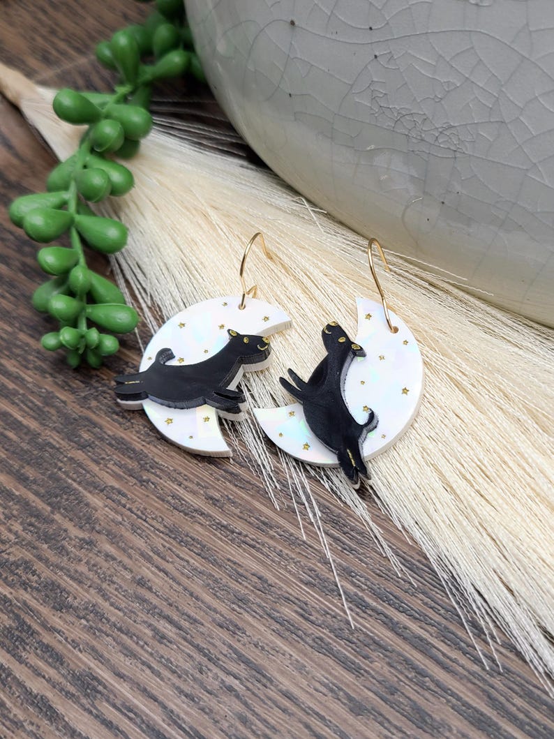 May include: A pair of dangle earrings featuring a white crescent moon with a subtle iridescent finish and small gold star accents. A black dog silhouette is layered on top of each moon, and the earrings hang from gold-toned metal ear wires.