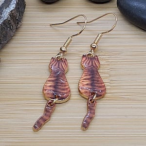 May include: A pair of gold-colored cat-shaped earrings. The earrings feature a brown and tan striped design on the cat's body and tail. The earrings are hanging from gold-colored ear wires.