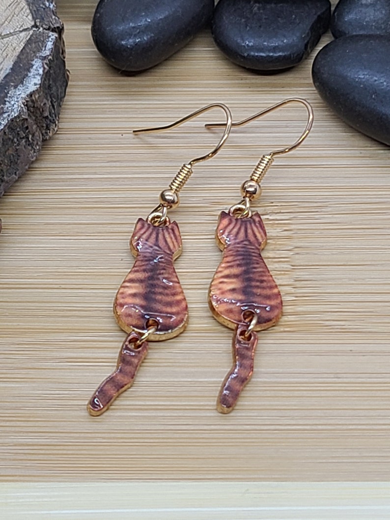 May include: A pair of gold-colored cat-shaped earrings. The earrings feature a brown and tan striped design on the cat's body and tail. The earrings are hanging from gold-colored ear wires.