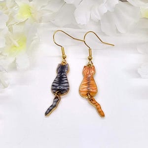 May include: A pair of gold-colored cat earrings. One earring features a gray and black striped cat, while the other showcases an orange and brown tabby cat. The earrings are suspended from gold-colored hooks.