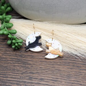 May include: A pair of dangle earrings featuring white crescent moon charms decorated with small gold stars. One earring displays a black cat silhouette, while the other features a tan cat silhouette. Both charms hang from thin gold-toned metal hooks.
