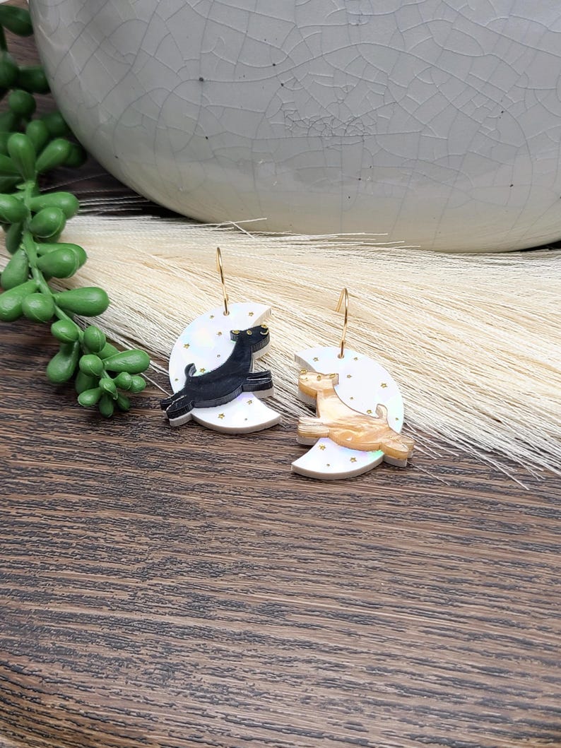 May include: A pair of dangle earrings featuring white crescent moon charms decorated with small gold stars. One earring displays a black cat silhouette, while the other features a tan cat silhouette. Both charms hang from thin gold-toned metal hooks.