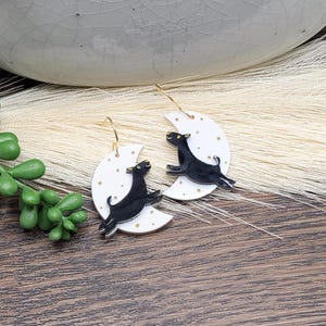 May include: A pair of dangle earrings featuring white crescent moon charms decorated with small gold stars. Each moon has a black dog silhouette leaping across the surface. The earrings are attached to thin gold-toned metal ear wires.