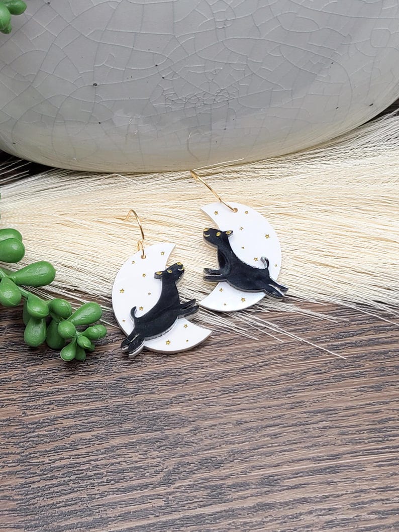 May include: A pair of dangle earrings featuring white crescent moon charms decorated with small gold stars. Each moon has a black dog silhouette leaping across the surface. The earrings are attached to thin gold-toned metal ear wires.