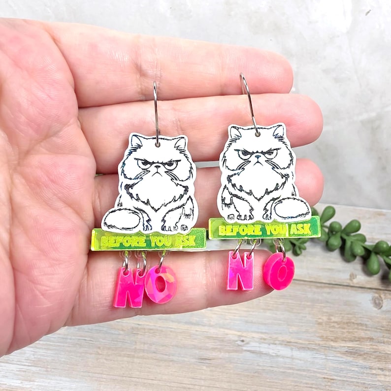 May include: A pair of dangle earrings featuring a white, grumpy cat illustration with the text "BEFORE YOU ASK". Below the cat, pink letters spell "NO". The earrings have silver-colored hooks.